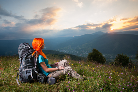 Back view of female backpacker with a backpack sitting in the grass with wild flowers on a hill and looking over the beautiful mountains, village in the valley and incredible cloudy sky at sunsetの写真素材