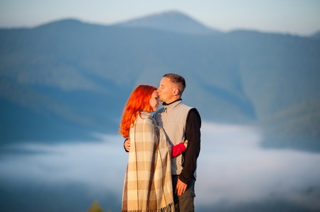 Romantic couple tourists kissing and hugging each other against beautiful mountain landscape with morning haze over the mountains on background. Red-haired girl covered with a blanket. Close-upの写真素材