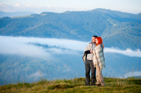 Romantic couple covered with a plaid standing together on a hill, enjoying beautiful mountain landscape with morning haze over the mountains and forestsの写真素材