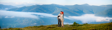 Sweety couple hikers covered with a blanket standing together on a hill, enjoying beautiful mountain landscape with morning haze over the mountains and forests. Panoramaの写真素材