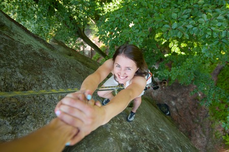 Young happy female climber hanging on two hands on high boulder. Man giving a helping hand to reach a peak of mountainの写真素材