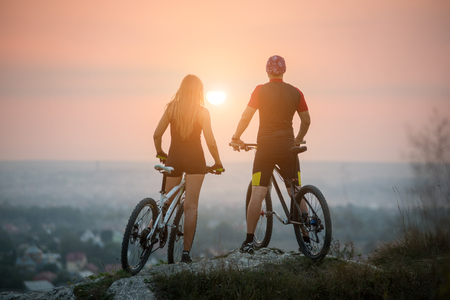 Rear view of man and woman with long hair on the mountain bikes enjoying the sunset. Bright sun in between. Blurred backgroundの写真素材