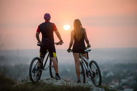 Rear view of guy and girl cyclists on the sports bikes enjoying the beautiful sunset. Bright sun in between. Blurred background.の写真素材