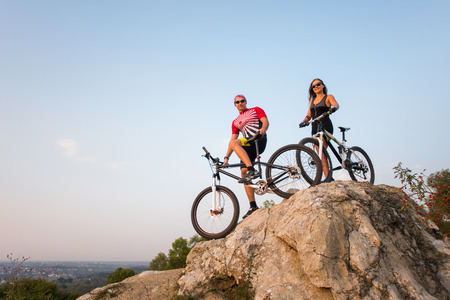 Cyclists in sunglasses stay on the bicycles at the precipice of hill against evening sky and looking to the camera. Bottom view. Pink Kinesio tape glued on the girl's hand.の写真素材