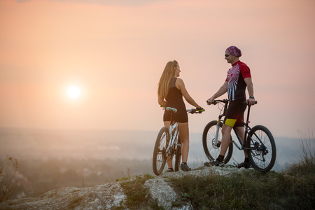 Rear view cyclist couple with mountain bikes standing on a rock, enjoying the evening scenery, looking to each other. Blurred backgroundの写真素材