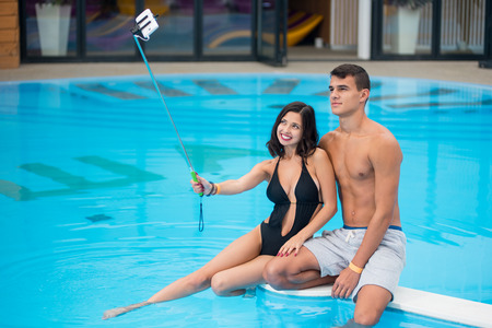 Young couple sitting on the edge of the swimming pool with perfect aqua water and taking selfie photo on the phone with selfie stick on the luxury resortの写真素材