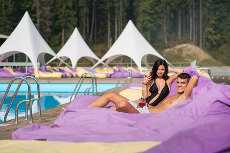 Happy couple - man and woman relaxing near swimming pool on cushioned loungers with drinks at the luxury resort on the blurry background of forest of the Carpathiansの写真素材
