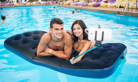 Close-up of young smiling couple making selfie photo on the phone with selfie stick on mattress in pool and on their summer vacation.の写真素材