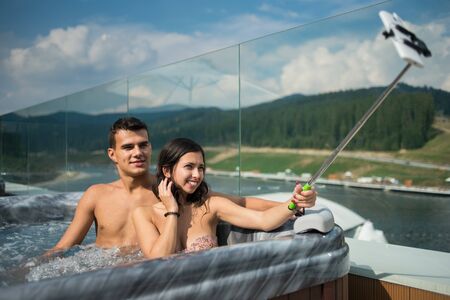 Romantic couple enjoying hot tub outdoors and making selfie photo on the phone with selfie stick against blurred background of natureの写真素材