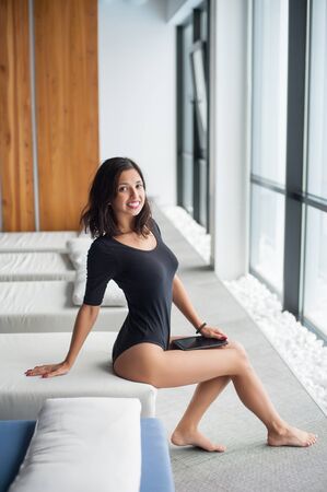 Smiling young woman in black bodysuit enjoying on white lounger near big windows and holding tablet computer in hands at lounge zone in the spa resortの写真素材