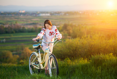 Brunette woman in Ukrainian embroidery walking with white bicycle at hill on a blurred background of greenery and small townの写真素材