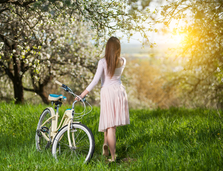 Graceful woman in a white dress standing with her back and holding a bicycle in fresh green grass under flowering trees in the blooming gardenの写真素材
