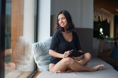 Joyful woman in black bodysuit sitting cross-legged holding in hands tablet computer and looks in the window at lounge zone in the luxury resortの写真素材