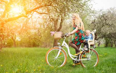 Beautiful female with long blonde hair in dress riding vintage bicycle with baby in bicycle chair, looking to the camera. In the basket lay a bouquet of lilacs, against spring gardenの写真素材