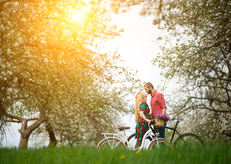 Loving young couple with bikes kissing against the background of blooming trees and fresh greenery in spring garden. Couple together enjoying romantic holidays. Side viewの写真素材