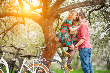 Romantic couple kissing near the tree and bicycles in spring garden. Woman with long blond hair in flowered dress sitting on tree branch and man stand near her. Side viewの写真素材