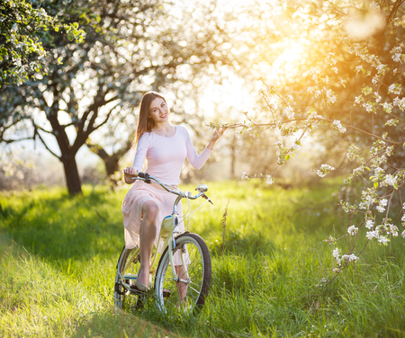 Young girl with long brunette hair wearing light dress riding a retro white bike holds a hand a branch of a blossoming tree on the blurred background of spring garden.の写真素材