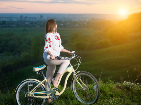 Young female in Ukrainian embroidery holding vintage bicycle at hill and looking into the distance, on a blurred background of greenery and small town at sunsetの写真素材