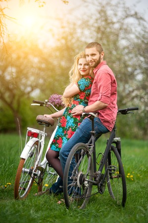 Two smiling lovers standing with bicycles and looking to the camera in the spring garden, against the background of trees and fresh greeneryの写真素材