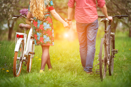 Young couple walking with bicycles holding hands back to camera, in the spring garden, against the background of trees and fresh greenery. Close-upの写真素材