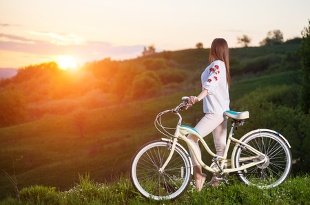 Woman in Ukrainian embroidery with bicycle standing at hill and looks at beautiful sunset with a blurred background of greeneryの写真素材