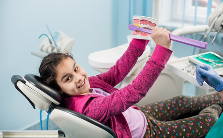 Smiling girl in dentist chair showing proper tooth-brushing using dental jaw model and big toothbrush in dental clinic. Dentistry, early prevention, oral hygiene conceptの写真素材