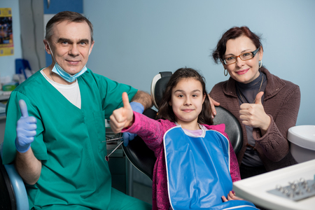Portrait of pediatric dentist and young girl with her mother on the first dental visit at the dental office. People is smiling and showing thumbs up. Dentistry, medicine and health care conceptの写真素材
