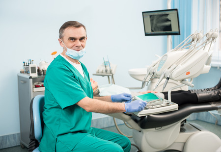 Handsome male dentist with dental tools in the dental office. Doctor wearing mask, green uniform and blue gloves. On the background patient and monitor with X-ray the patient's teeth. Dentistryの写真素材