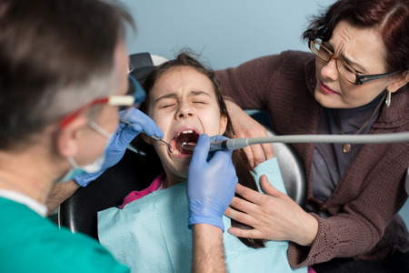 Young girl with her mother on the first dental visit. Senior pediatric dentist treating patient girl teeth at the dental office. Medicine, stomatology and health care concept. Dental equipmentの写真素材