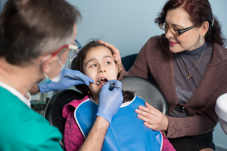 Young girl with mother on the first dental visit. Senior pediatric dentist doing first check-up for patient at the dental office. Dentistry, medicine, stomatology and health care concept. Dental equipmentの写真素材