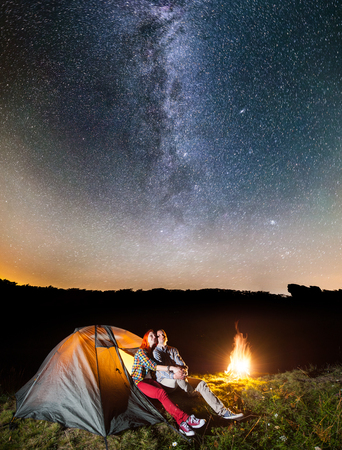 Happy couple tourists looking to the shines starry sky, sitting near glowing tent and campfire in the camping at night. Astrophotographyの写真素材