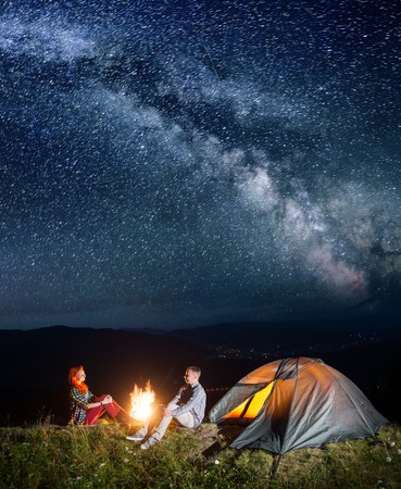 Night camping. Happy couple backpackers sitting by bonfire and tent under incredibly beautiful starry sky. Village in the valley on the background. Long exposureの写真素材