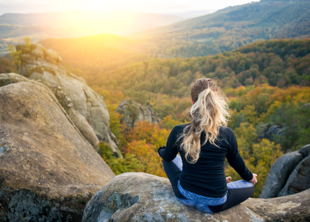 Young fit girl is practicing yoga and doing asana Siddhasana on the top of the high rocky mountain in the evening. Beautiful sunset, autumn forests, rocks and hills on the backgroundの写真素材