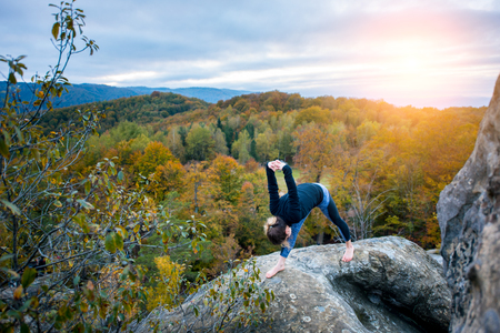 Sporty fit girl is practicing yoga on the top of the high rock in the evening. Autumn forests, rocks and hills on the backgroundの写真素材