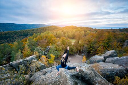 Flexible fit woman is practicing yoga and doing asana Virabhadrasana 1 on the top of the huge boulder in the evening. Autumn forests, rocks and hills on the backgroundの写真素材
