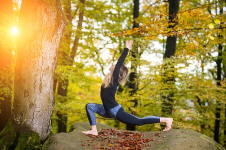 Beautiful young woman is practicing yoga and doing asana Virabhadrasana 1 - Beautiful young woman is practicing yoga in autumn forest on the big stoneの写真素材