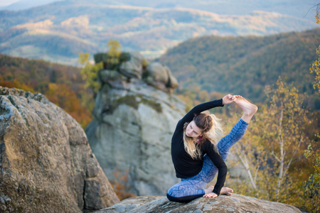 Flexible young woman is practicing yoga and doing asana Surya Yantrasana on the top of the high rocky mountain. Autumn forests, rocks and hills on the backgroundの写真素材