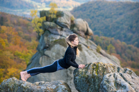 Peaceful fit woman is practicing yoga and doing asana Urdhva Mukha Shvanasana on the top of the mountain. Autumn forests, rocks and hills on the backgroundの写真素材