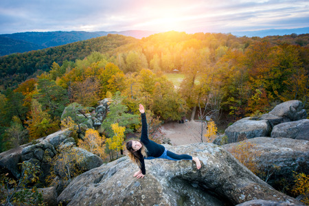 Pretty young female is practicing yoga on the top of the mountain in the evening. Beautiful sunset, autumn forests, rocks and hills on the backgroundの写真素材