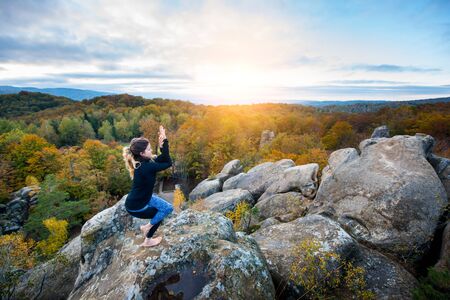 Attractive young female is practicing yoga and doing asana Garudasuna on the top of the huge boulder in the evening. Beautiful sunset, autumn forests, rocks and hills on the backgroundの写真素材