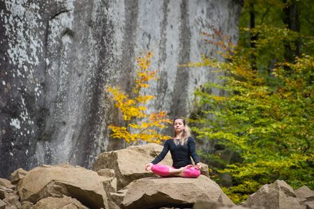 Pretty flexible woman is practicing yoga and doing asana Siddhasana on the boulder in the nature. Huge rocks on the backgroundの写真素材