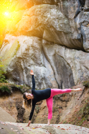 Sporty fit girl is practicing yoga and doing asana Ardha Chandrasana on the boulder in the nature. Huge rocks on the backgroundの写真素材