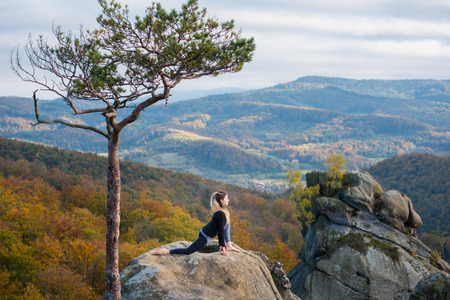 Active fit woman is practicing yoga on the top of the mountain near big tree. Autumn forests, rocks and hills on the backgroundの写真素材