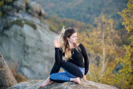 Calm young woman is practicing yoga and doing asana Arha Matsyendrasana on the top of the high rocky mountain in the evening. Autumn forests, rocks and hills on the backgroundの写真素材