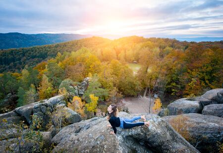 Pretty young woman is practicing yoga and doing asana Urdhva Mukha Shvanasana on the top of the mountain in the evening. Beautiful sunset, autumn forests, rocks and hills on the backgroundの写真素材