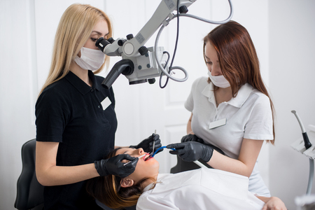 Two female dentists checking up patient teeth with dental tools - microscope, mirror and probe at dental clinic office. Medicine, dentistry and health care concept. Dental equipmentの写真素材