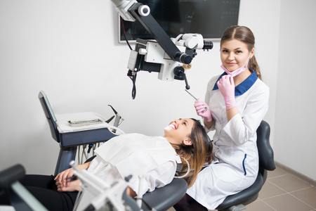 Attractive female dentist with happy woman patient during treatment with microscope in modern dental office. Doctor looking to the camera, holding dental tools. Dentistry. Stomatology equipmentの写真素材