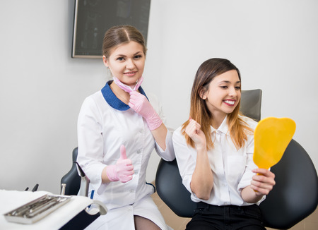 Young happy female patient checking their teeth at mirror after dental procedure in dental office. Female dentist looking to the camera and showing thumbs up. Dentistryの写真素材