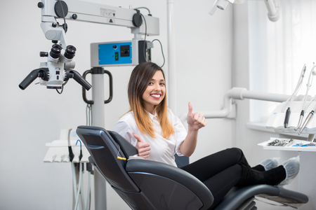 Happy female patient with perfect white teeth sitting in dental chair, smiling and showing thumbs up after treatment at modern dental clinic. Dentistry, medicine and healthcare concept.の写真素材