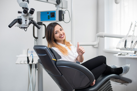 Beautiful female patient with perfect white teeth sitting in dental chair, smiling and showing thumbs up after treatment at modern dental clinic. Dentistry, medicine and healthcare concept.の写真素材